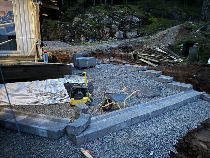 Byggarbetsplats med markduk, lecablock och grus under arbete för grunden. En skottkärra och en komprimeringsplatta syns för förberedelser.