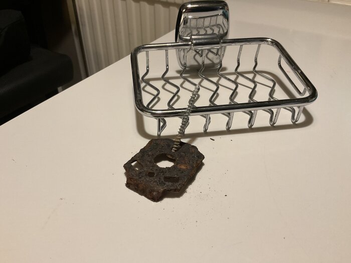 En trasig kromad duschkorg med en rostig väggplatta och skruv ligger på en vit yta.