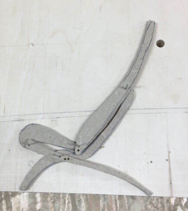 Skiss av en liggande stol, konstruerad av grå pappbitar sammanfogade med små hål, placerad på en vit bakgrund.