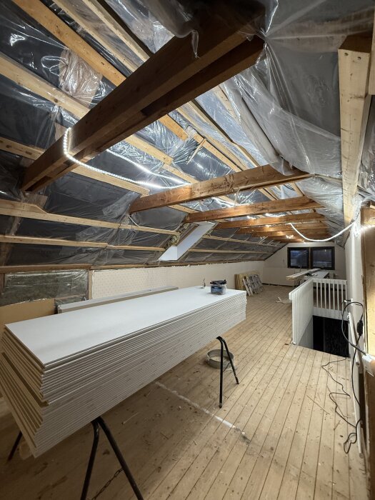 Orenoverat vindsutrymme med synliga träbalkar och staplade gipsskivor, plast på taket och omålat trägolv under pågående renovering.