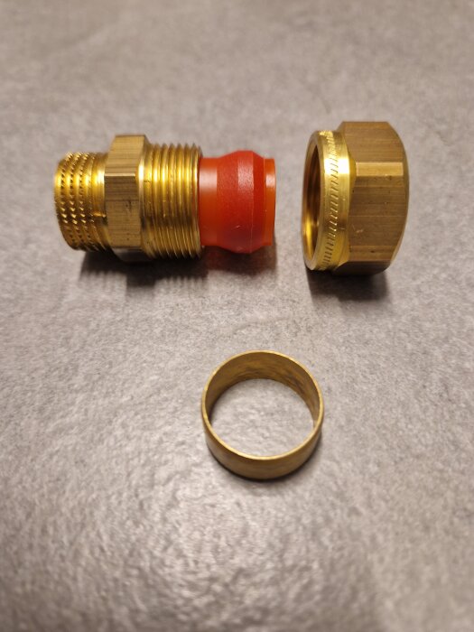 Kopplingsdetaljer med en klämringskoppling och rörgänga av mässing samt en röd tätning, mot en grå bakgrund.