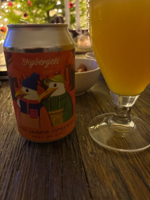 Burk med öl märkt Stigbergets, "After Laughter Comes Beers Hazy IPA 6.5%", bredvid ett glas öl på ett träbord, med julgran i bakgrunden.