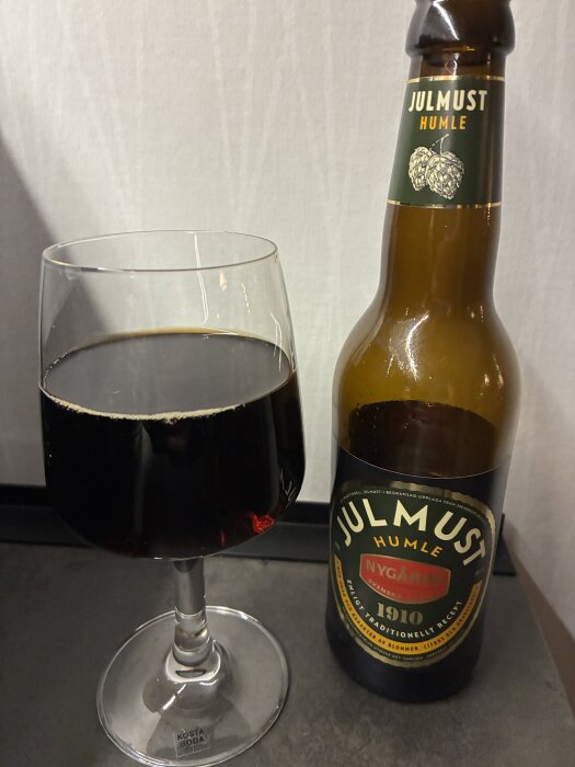 Ett vinglas med mörk dryck bredvid en flaska julmust humle från Nygårda med etikett.