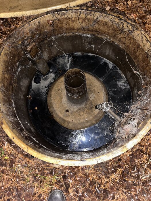 Bild av ett öppet betonghål med metallrör och avlagringar, omgiven av jord och barr. Fråga om identifiering.