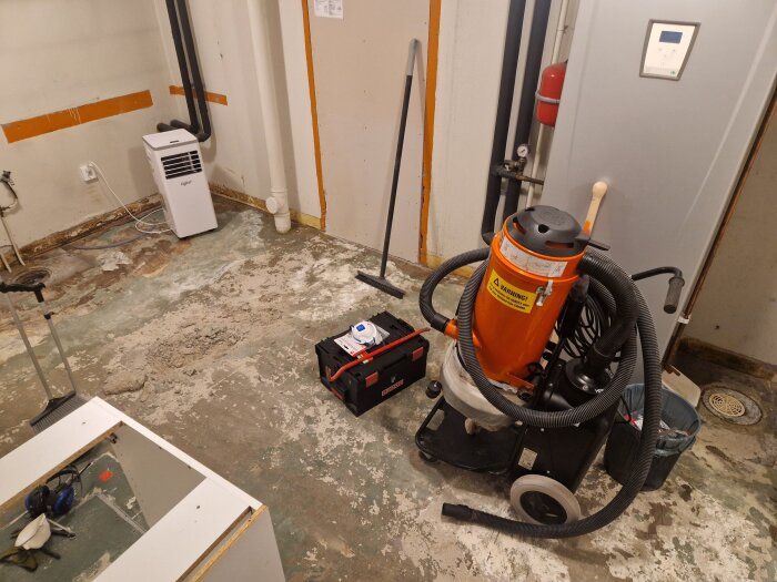 Rum med renoveringsutrustning, betonggolv och två gjutjärnsbrunnar. En orange dammsugare och en verktygslåda syns också.