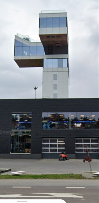 Modern byggnad med svart fasad och glaspartier. Över byggnaden finns ett fackverksliknande torn med utstickande del i glas.