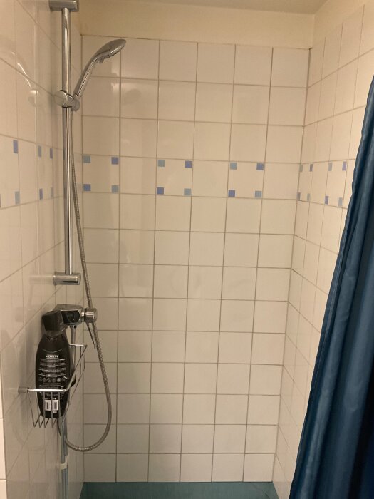 Vitkaklad dusch med duschmunstycke och blå duschdraperi. Spår av rost på kaklet, omfogad. Flaskor i metallkorg.