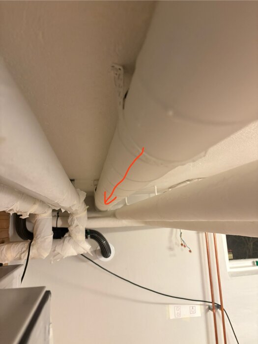 Rörinstallationer i tvättstuga med spirorör längs taket och en röd pil som visar ventilationsriktningen. Ytterligare rör och kablar syns.