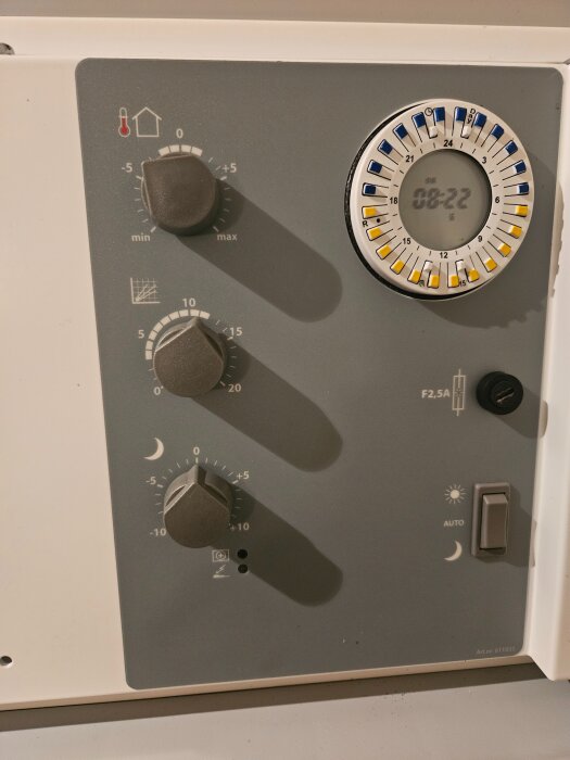 Grå panel med tre vred för temperaturjustering och en timerklocka. En strömbrytare med symboler för sol och måne finns till höger.