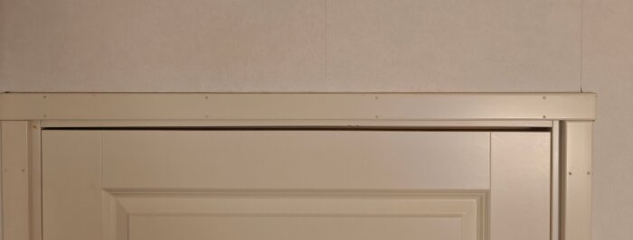 Övre delen av en vit dörrkarm mot en ljus vägg med synliga skruvar längs karmens kanter.