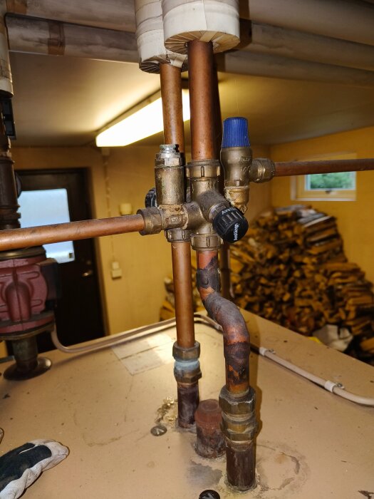 Rörsystem ovanför en vedpanna med flera ventiler och ett blått vred, omgivet av kopparrör och en hög med ved i bakgrunden.