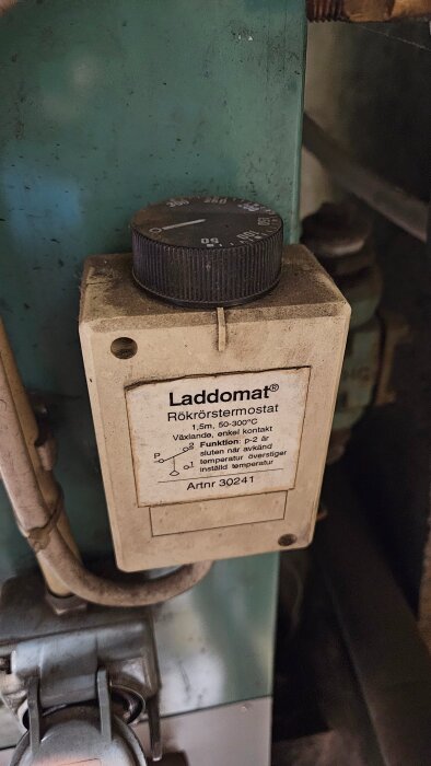 Laddomat rökrörstermostat installerad på en grön pannvägg med en ratt för temperaturjustering och etikett som beskriver funktion och artikelnummer.