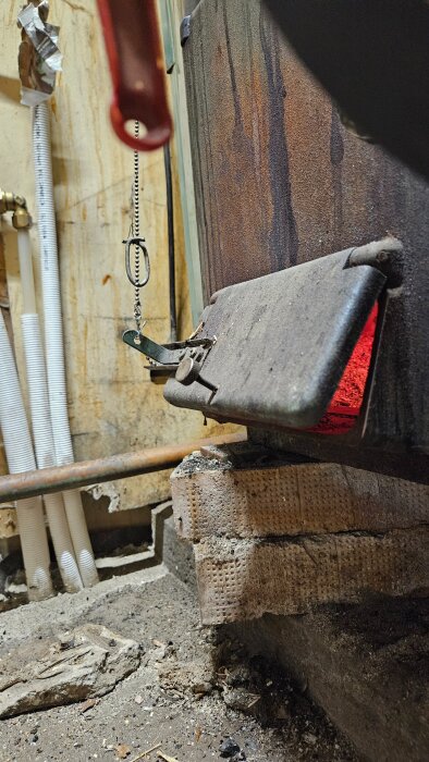 Bild på en sotig lucka med kedjereglering, del av en eldstad. Luckan är delvis öppen och inuti syns glödande aska.