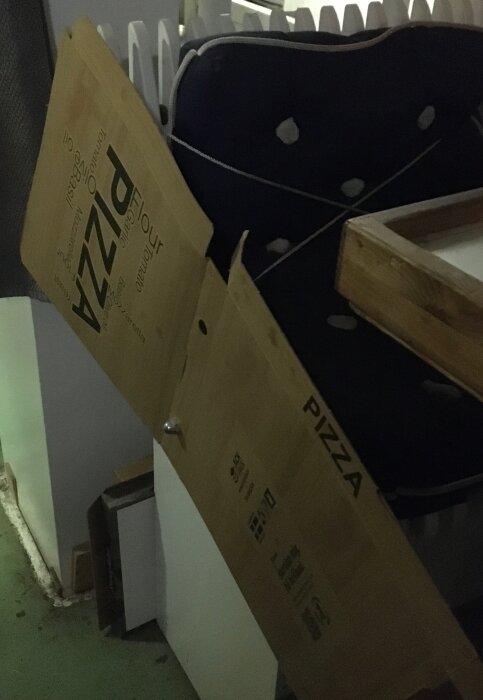 Pizzakartong lutad mot en stol med blå dyna, synlig text på kartongen med ordet "Pizza". Bakgrunden visar en vägg och golv.