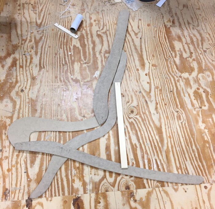 Utspridda träbitar och kartongmallar på ett plywoodgolv, troligen för ett träprojekt, med en pappersrulle i övre delen av bilden.
