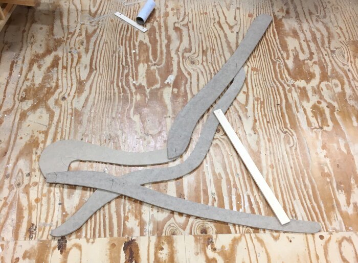 Golv av plywood med utskurna former av kartong och en träbit som stöd, omgivet av verktyg och material.