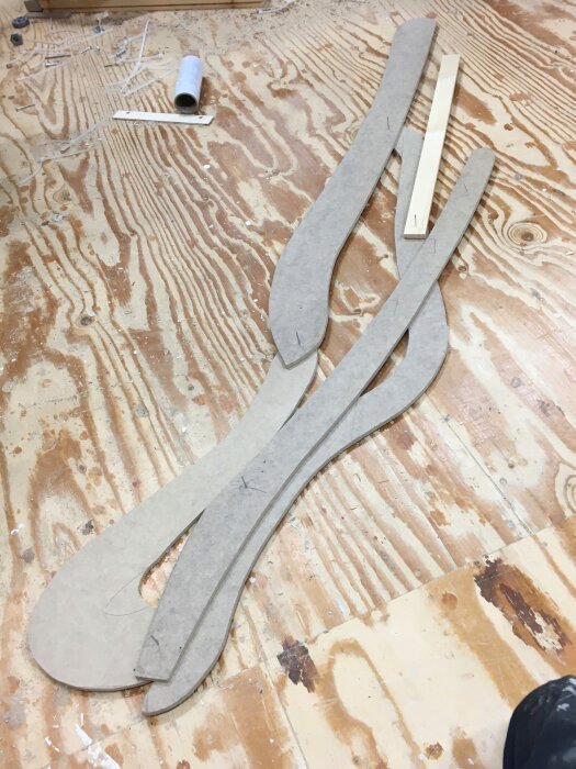 Formade träbitar och kartongmallar ligger på ett trägolv i en verkstad, tillsammans med en tom kartongrulle och träspånor.