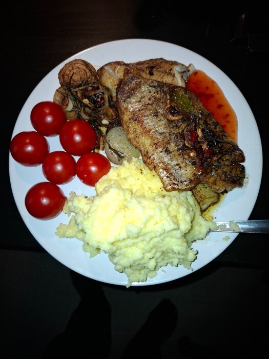 En tallrik med stekt kolja, mosade potatisar, körsbärstomater och stekt lök, samt en sås vid sidan.