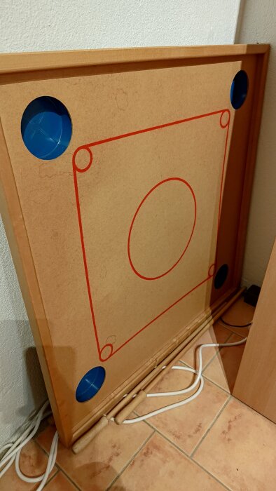 Gammalt spelbräde hittat på vind, med matta yta och röda markeringar, placerat mot en vägg. Fyra blå hål i hörnen för spelpjäser.