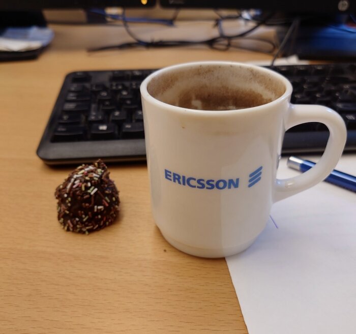 En Ericsson-mugg med kaffe står på ett skrivbord bredvid en chokladboll med strössel, framför ett tangentbord och ett papper.