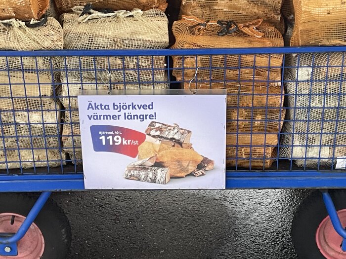 Skylt med texten "Äkta björkved värmer längre!" framför staplade säckar med ved, pris 119 kr för 40 L.