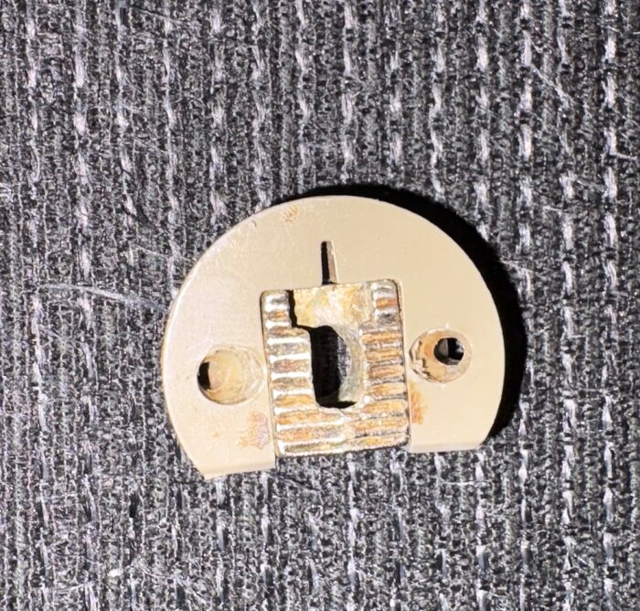 Skruvplatta i metall med två monteringshål och slitna kanter, placerad på en mörk tygbakgrund.