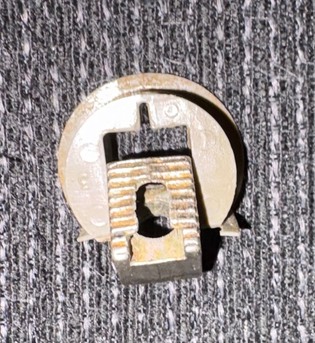 Rund metallkoppling med fyrkantigt mittparti, liggande på en grå texturerad yta.