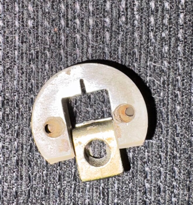 Metallfäste med två skruvhål på en grå, texturerad yta.