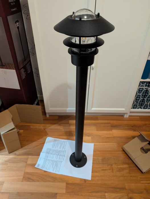 Svart utomhuslampa på stång står på ett trägolv, omgiven av kartonger och manualpapper. Lampan har en modern design med glaskupa.