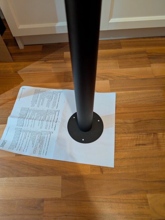 Svart metallben monterat på ett trägolv, med en rund basplatta ovanpå ett papper med text.