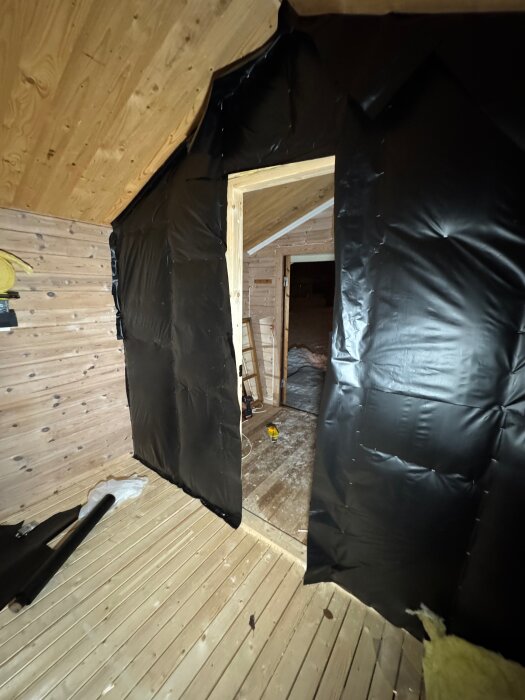 Svart fuktspärrfolie monterad på isolerad innervägg i en blivande bastu, trägolv och trätak synliga, öppning till annan del av friggeboden.