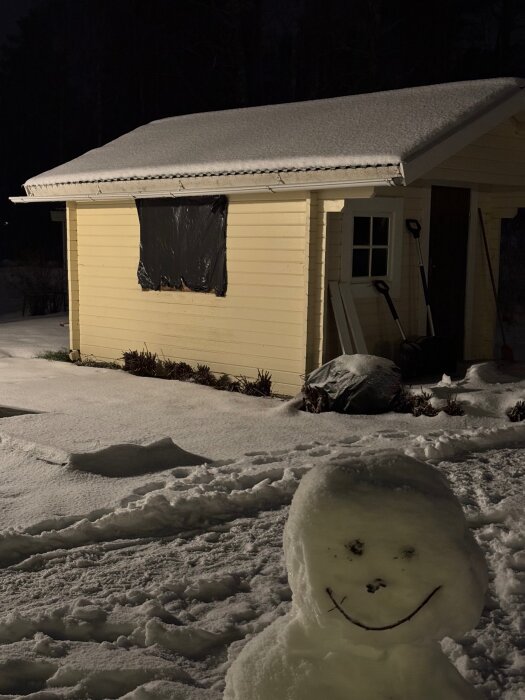 En snötäckta friggebod med plast över fönstret och en snögubbe i förgrunden på natten.