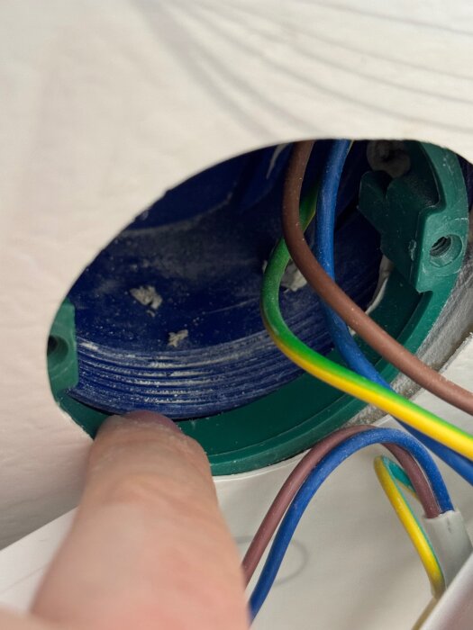 En närbild på olika färgade elkablar som löper genom ett runt väggfäste, med en person som pekar mot det inre av dosan.