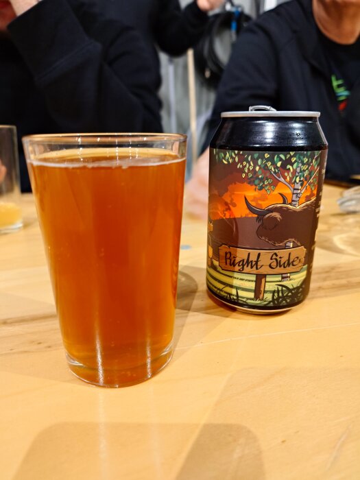 Ett glas med bärnstensfärgad öl står bredvid en färgglad ölburk på ett träbord.