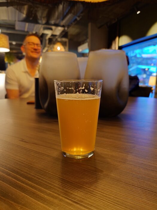Ett glas med ljus öl står på ett bord med oskarpa lampskärmar och en man i bakgrunden.