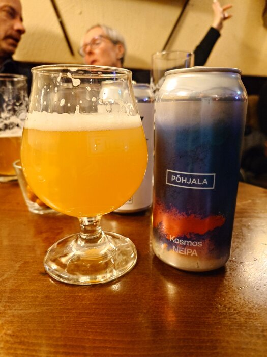 Ett glas med gul öl bredvid en ölburk med Põhjala-märket på ett träbord, personer sitter i bakgrunden.