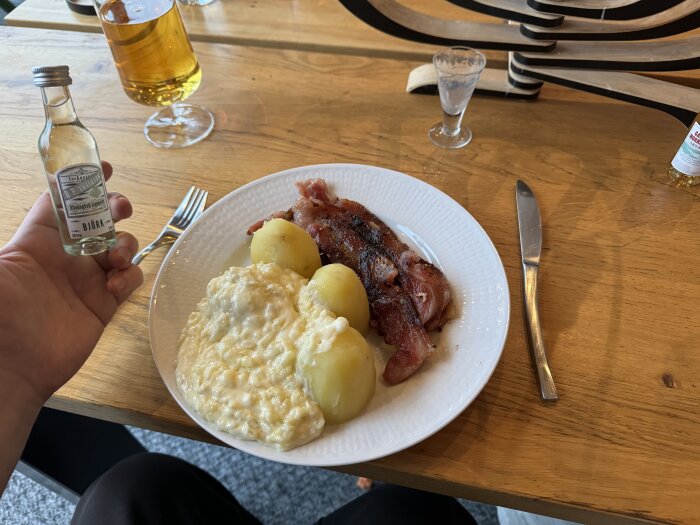 Tallrik med fläsk, potatis och mos på bord, liten flaska björksnaps i handen, glas med dryck, kaffe och snaps på bordet.