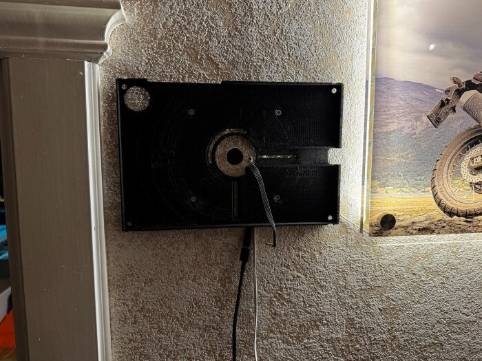 Svart väggfäste med sladdar monterat på en vägg bredvid en belyst tavla med motorcykelmotiv.