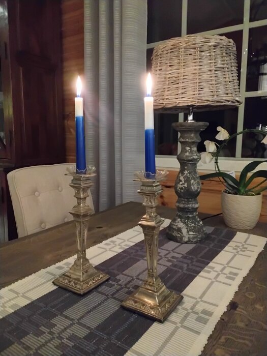 Två tända, blåvita ljus i silverljusstakar på ett bord med textilduk, bredvid en flätad bordslampa och en orkidé i kruka.