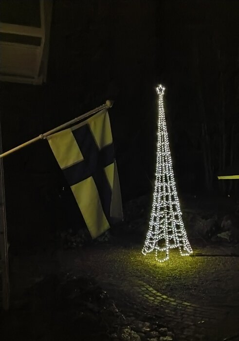Flagga med kors och julbelysning som formar ett träd i mörker.