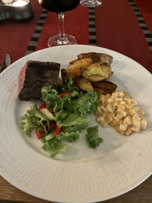Grillad rostbiff, Greek-style potatis, creamy corn och sallad på en tallrik med rödvinsglas i bakgrunden.
