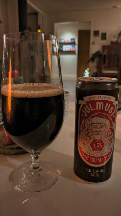 Ett glas fyllt med mörk julmust bredvid en burk märkt "Stark Julmust" med 4,5% alkohol, placerad på ett bord i ett inomhusmiljö.
