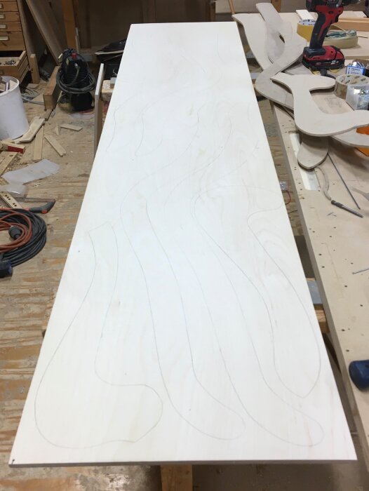 Plywoodskiva på arbetsbänk i verkstad, konturer av formen tecknade. Verktyg och material synliga i bakgrunden.