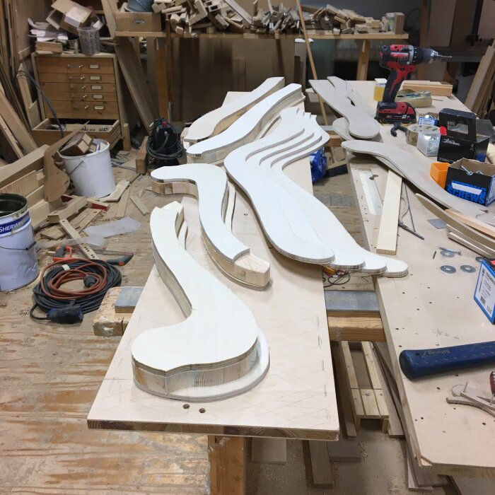 Utskurna träbitar i olika former ligger på ett bord i en verkstad med verktyg och byggmaterial runtom.