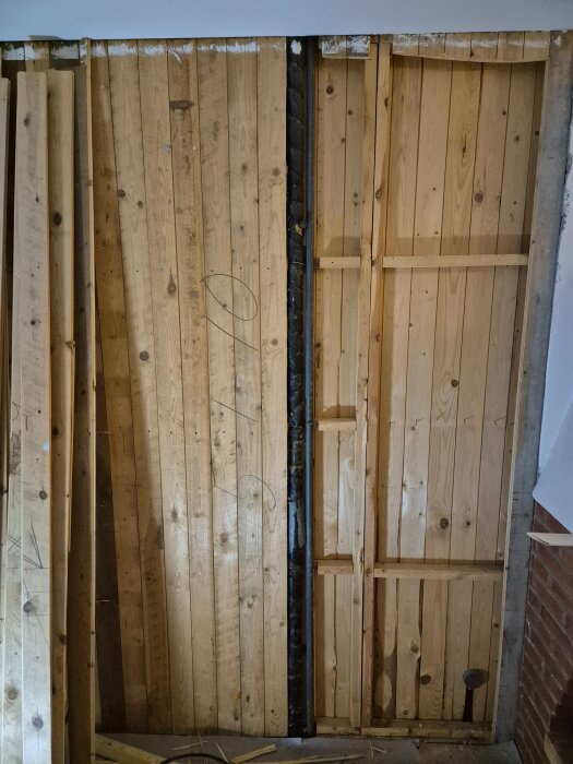 Två lodräta rör syns bakom en brun trävägg. Det ena röret är delvis isolerat och ser varmförzinkat ut. Runt rören ses träplankor och en del av en tegelvägg.