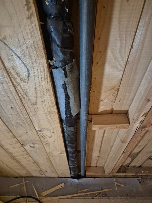 Lodräta rör bakom träpanel, ett med svart isolering och ett metalliskt, sträcker sig ner till golv; del av ett väggprojekt i renovering.