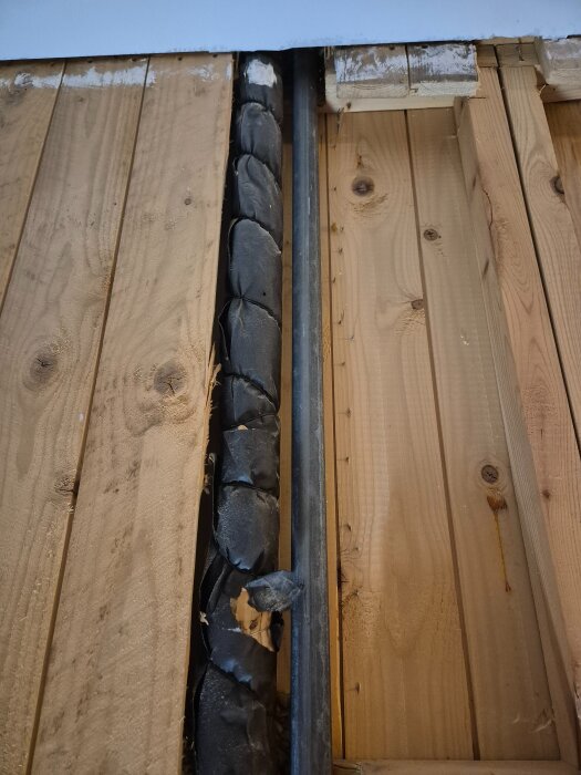 Två lodräta rör bakom träplankor, ett av rören är isolerat med svart material och det andra är varmförzinkat.