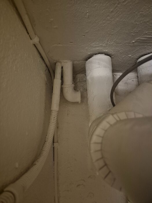 Vita lodräta rör av osäker funktion syns i ett hörn inomhus. Rören går genom taket och ett är delvis isolerat. En kabel löper längs väggen.