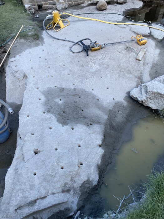 Bergarbetesplats med borrhål i berget, omgiven av verktyg och vattenpölar.