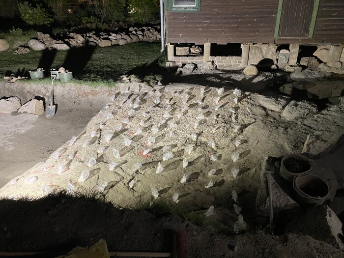 Berg med borrhål täckta av plastpåsar nära ett hus under renovering, med skugga och redskap placerade omkring.